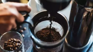 A person pressing down on a coffee plunger, with the grounds at the bottom of the container and the freshly brewed coffee being poured into a cup.