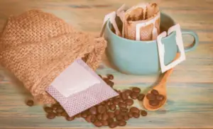 Drip coffee bag inside of a coffee cup with an open bag of fresh coffee beans beside the cup.