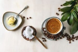 Bulletproof coffee in a black ceramic mug. To the side is a spoon of ghee butter and a spoon of coconut milk.