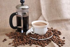 Coffee beans scattered around a cup of coffee in a white cup with a french press full of coffee in the background.