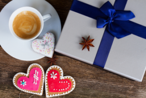 A cup of coffee with a heart shaped chocolate on the side, two heart shaped large cookies next to the cup coffee and a large beautifully wrapped gift to the side showing some of the best coffee gifts for valentine's day.
